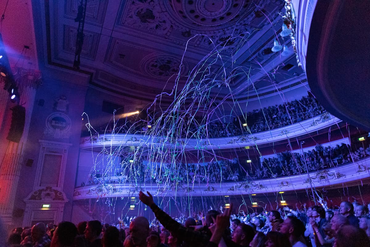 A lively crowd gathers in a large auditorium, illuminated by colorful lights and accompanied by vibrant music.