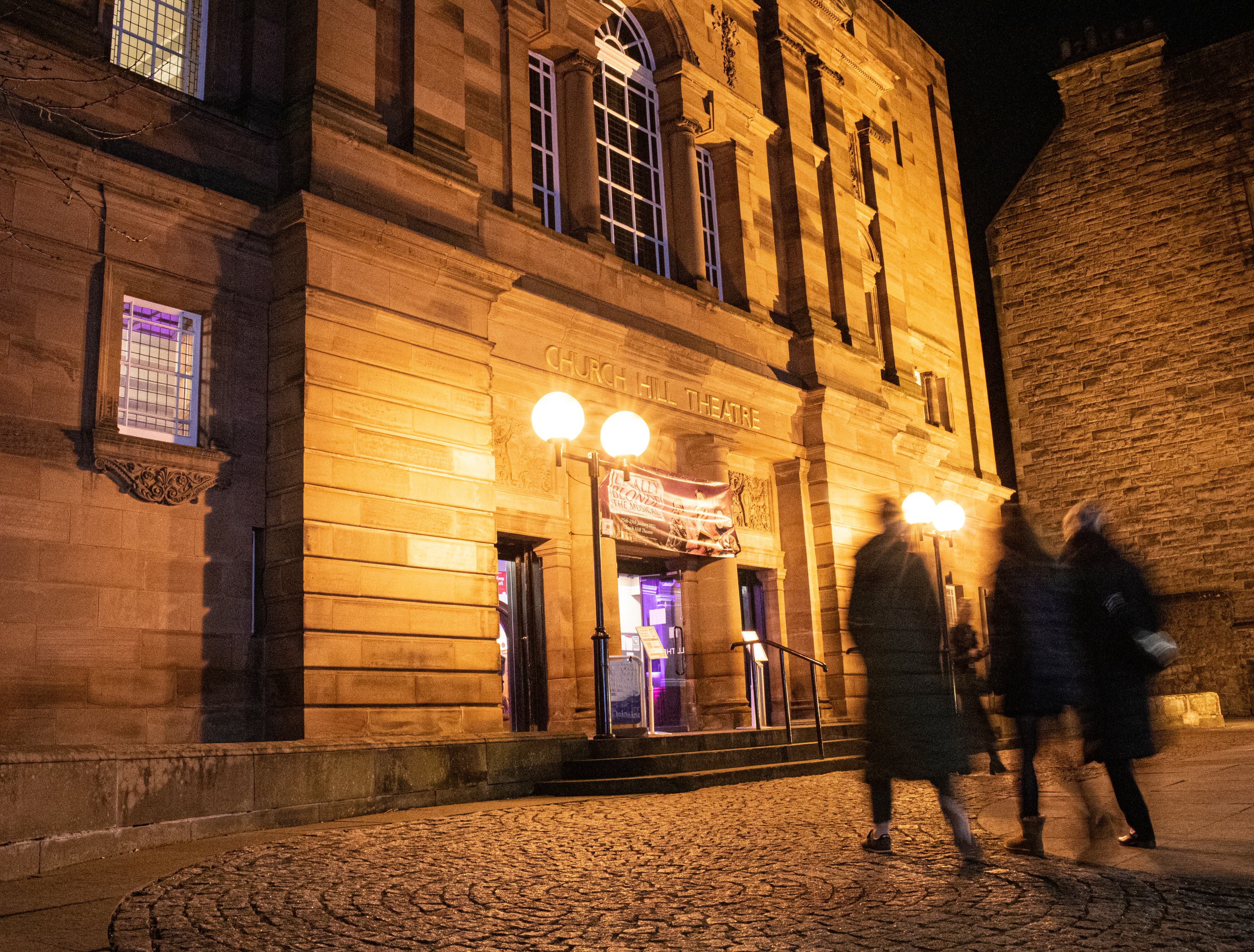 Nighttime scene of pedestrians entering a building, with warm lights illuminating the surroundings and the entrance door.