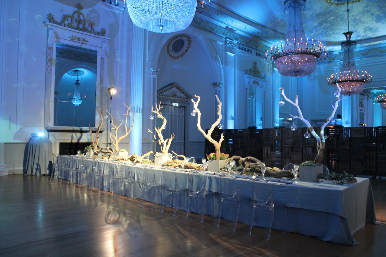 A long table covered with a tablecloth, elegantly set for dinner, under three large chandeliers. 