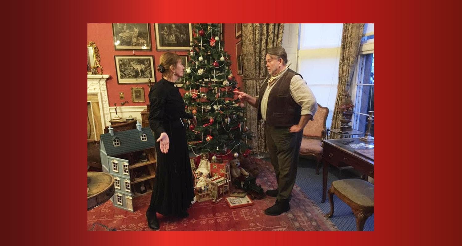 Two people in Edwardian costume stand beside a decorated Christmas tree in a cosy room in Lauriston Castle, surrounded by toys under the tree, a dolls house and framed art on the walls