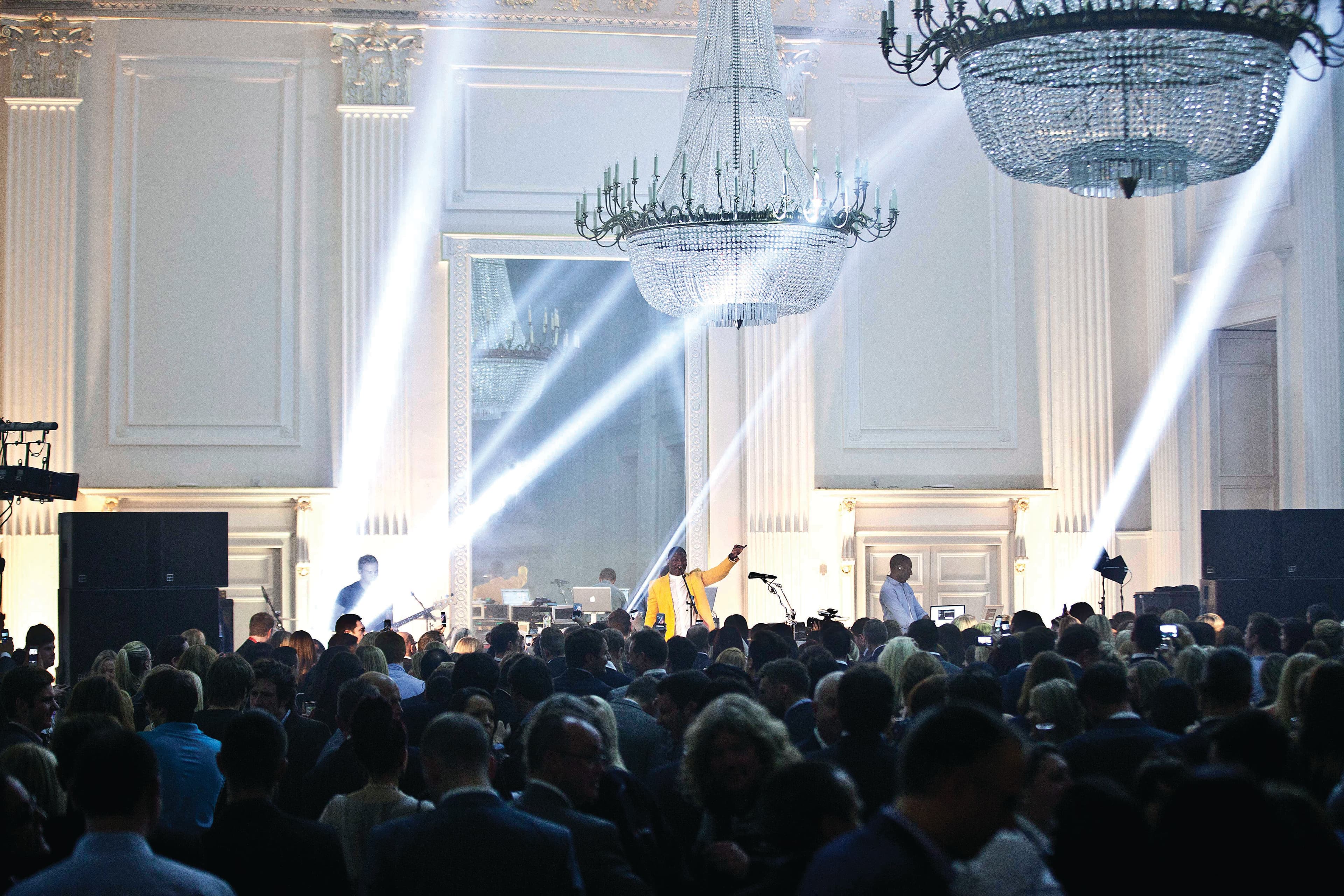 A bustling crowd fills a large room watching a concert, with people interacting and moving about. Bright spotlights shine over the crowd.