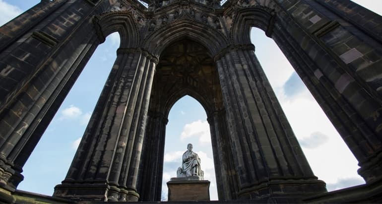 View from the bottom of the Scott Monument in Edinburgh. A marble statue of Sir Walter Scott is surrounded by tall stone pillars and a blue sky.