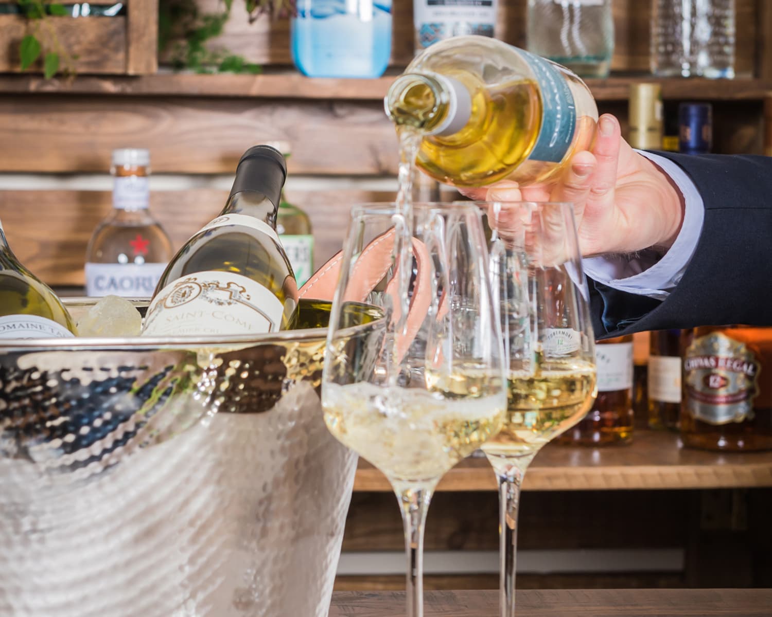 A hand pouring white wine into two glasses - a silver ice bucket is next to the glasses and a wine bar is in the background