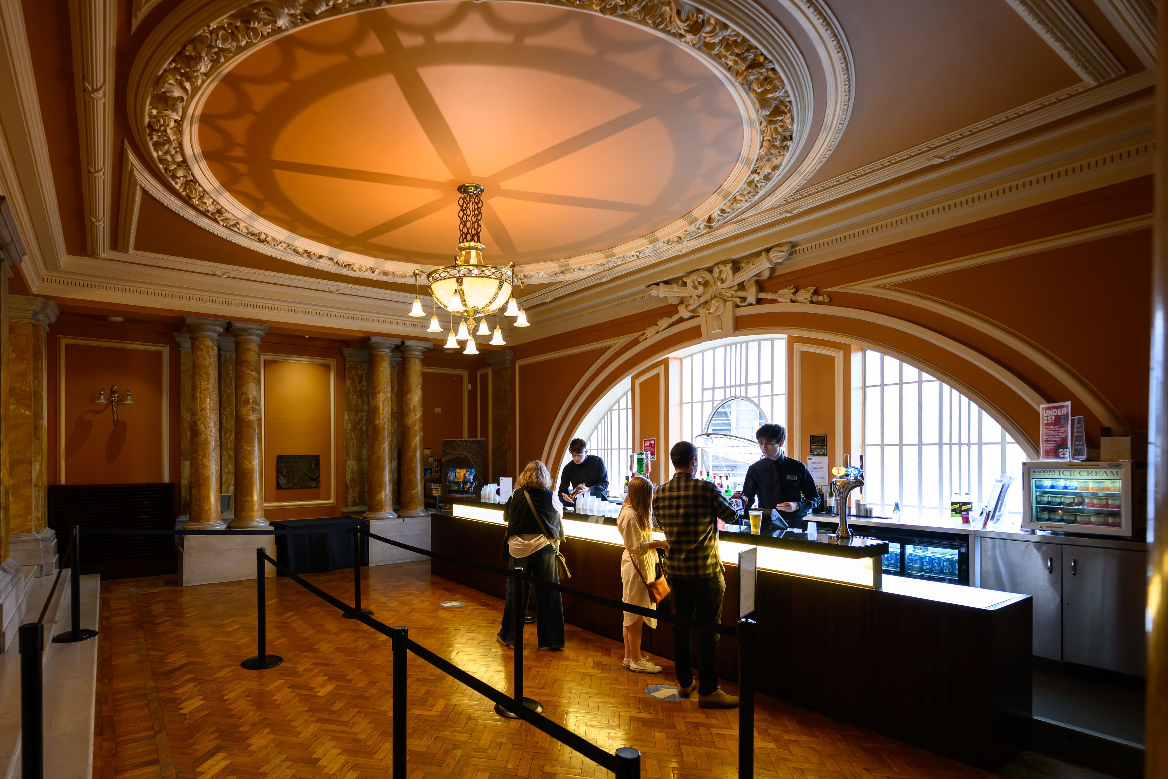 A group of people gathered around the bar. In front of a half circle window and a large architectural rose ceiling