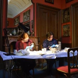 Two volunteers sit at a table in Edinburgh's Lauriston Castle cleaning and working with objects. The walls are full of paintings, and a large mirror is hung above a fireplace 