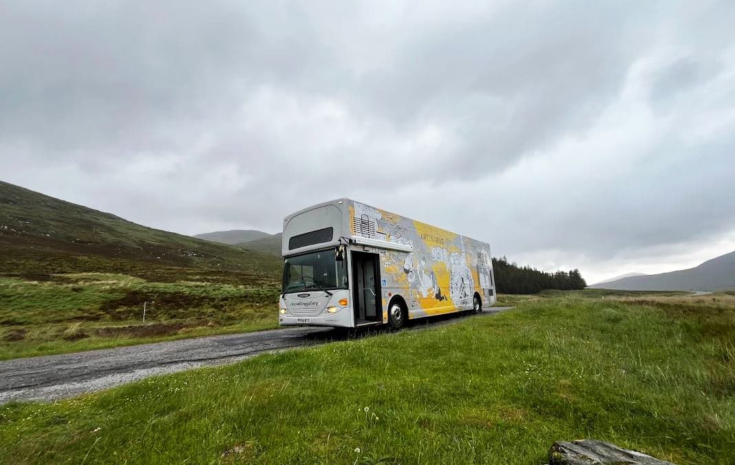 The travelling gallery bus is seen on a road in the middle of a field, emphasizing the rural landscape and open space around it.