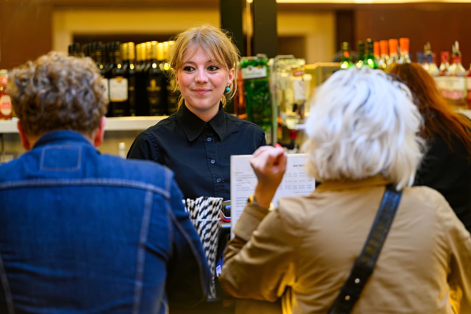 A woman member of staff at a bar, casually holding a drink, appears engaged in conversation with customers ordering drinks
