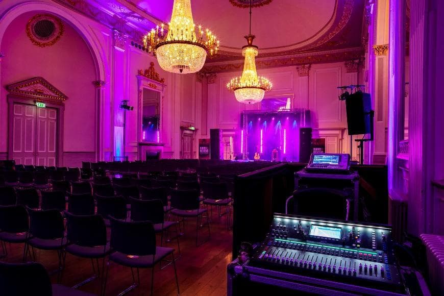 A spacious room in the Assembly Rooms in Edinburgh, illuminated with purple lighting, featuring a stage at the front for a performance or presentation. The lighting desk is in the foreground of the picture.
