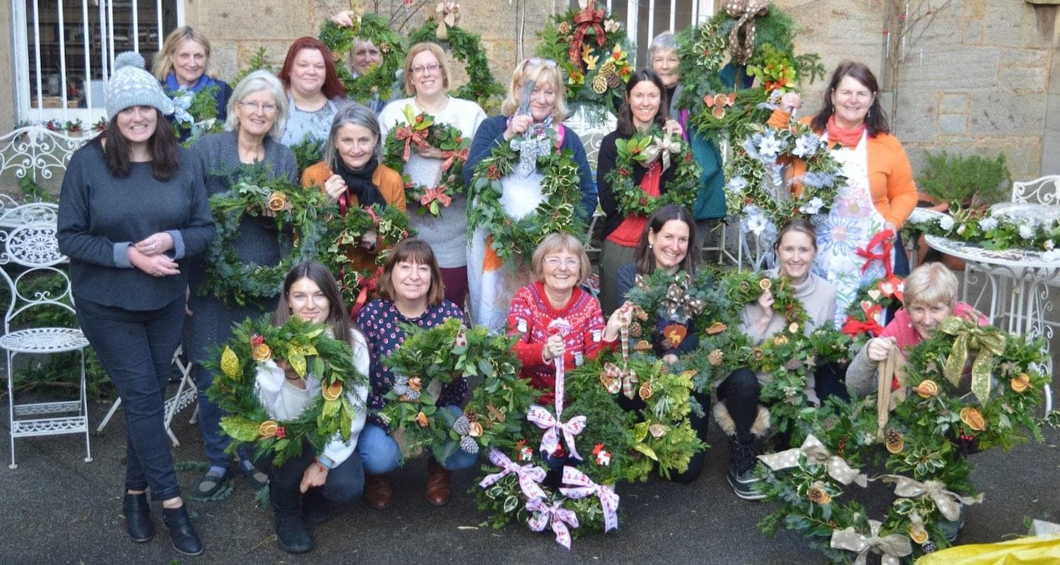 A group of people smile at the camera, each holding a Christmas wreath, decorated with ribbon, orange slices, pine cones, cinnamon sticks and wooden stars
