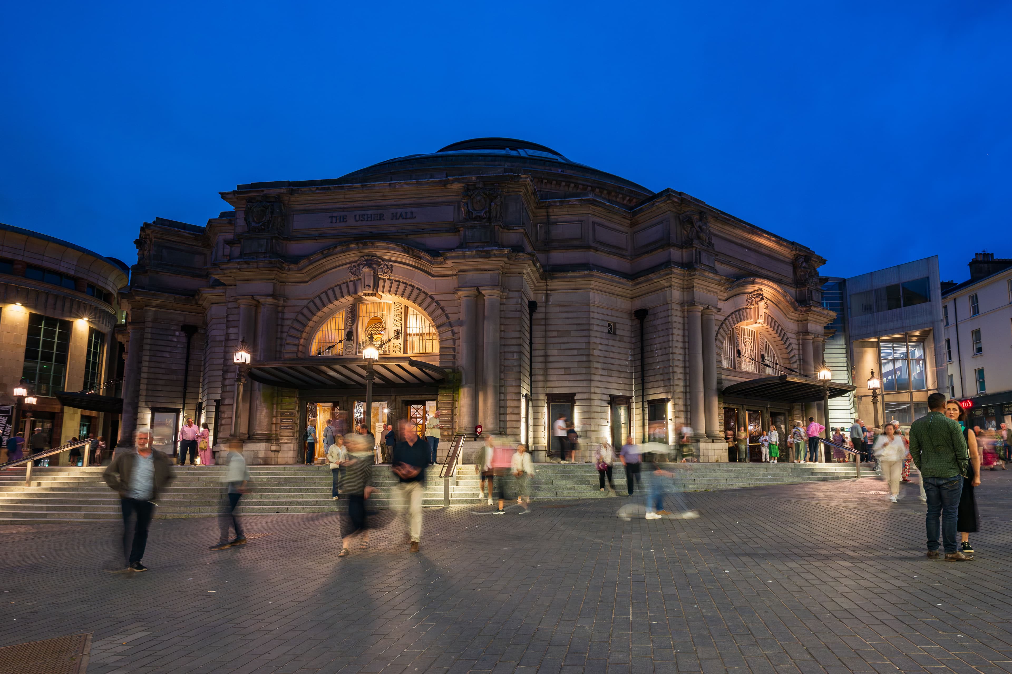 A Night time scene of people walking around the Usher Hall building, with warm lights highlighting its architecture.
