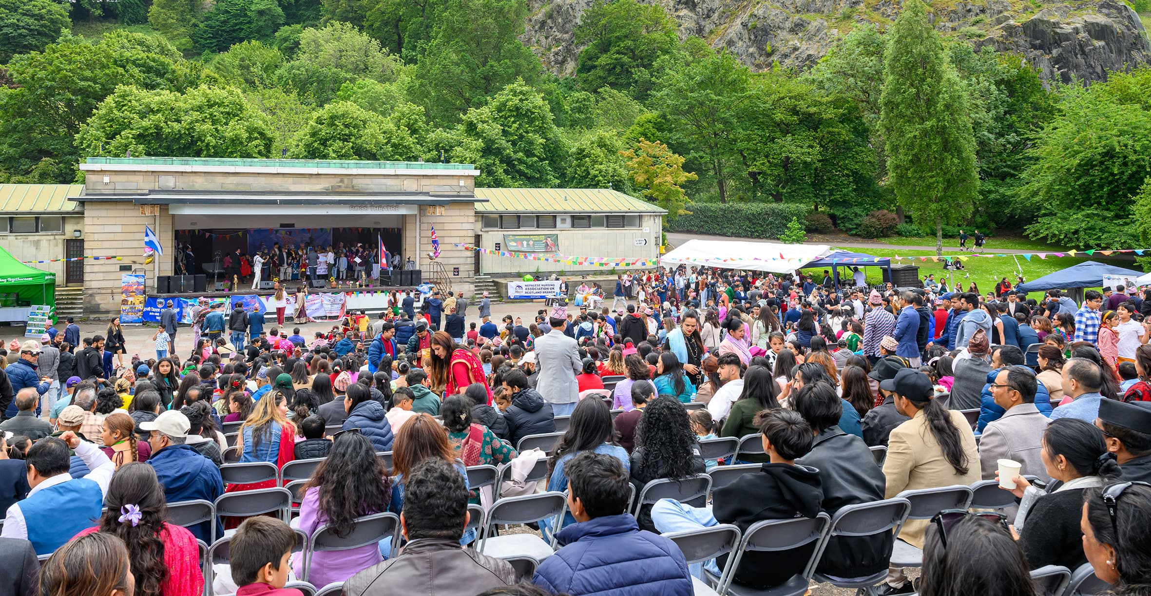 A vibrant scene of a large crowd in a park, with people mingling, sitting on the grass, and enjoying the atmosphere.